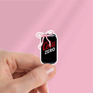 May include: A black Cola Zero can sticker with white text and a red logo, adorned with a pink bow. The sticker is held against a pink background.