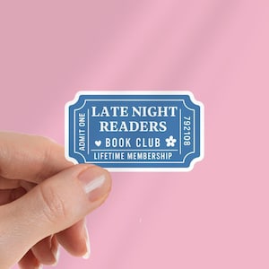 Può includere: Un adesivo blu e bianco a forma di biglietto. L'adesivo recita "LATE NIGHT READERS BOOK CLUB LIFETIME MEMBERSHIP" con una grafica a forma di fiore e cuore. L'adesivo è tenuto su uno sfondo rosa.