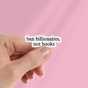 May include: A white sticker with the text "ban billionaires, not books" is held against a pink background. The sticker's text is in a black, sans-serif font. The image is a close-up shot.
