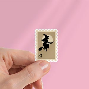 May include: A beige postage stamp with a black silhouette of a witch on a broomstick, flying in front of a full moon. The stamp has a white border and the number 31. The background is a solid pink color.