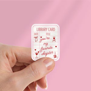 May include: A white sticker in the shape of a library card with red accents. The text "LIBRARY CARD" is at the top, with "DATE" and "TITLE" below. The sticker has the phrase "you're my favorite chapter" and illustrations of a bow, martini, and candle.