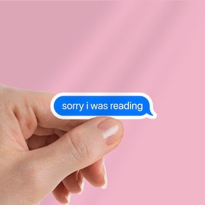 Könnte beinhalten: Ein Aufkleber in Form einer blauen Textblase mit weißem Rand, gehalten von einer Hand vor einem rosa Hintergrund. Der Aufkleber enthält den weißen Text: "sorry i was reading".
