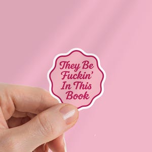 May include: A pink sticker with a scalloped edge and the text "They Be Fuckin' In This Book" in a cursive font. The sticker is held by a hand against a pink background.