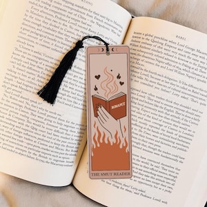 Peut inclure: Marque-page beige avec un dessin d'une main tenant un livre ouvert avec le mot "ROMANCE". Des flammes et des coeurs entourent le livre. Le texte "THE SMUT READER" est en bas. Une touffe noire est attachée.
