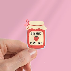 Op de afbeelding: Een sticker met een cartoon jam pot met de tekst "READING IS MY JAM" en een aardbei illustratie. De pot is rood met een crème deksel en een roze strik. De achtergrond is effen roze.