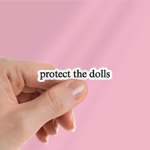 Puede incluir: Una pegatina blanca con el texto "protect the dolls" en negro, fuente sans-serif. La pegatina se sostiene entre los dedos sobre un fondo rosa.