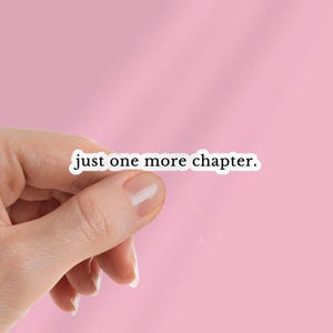 Puede incluir: Una pegatina blanca con el texto "just one more chapter." se sostiene sobre un fondo rosa. La pegatina tiene un borde negro y está en una fuente sans-serif. El fondo es de color rosa claro sólido.