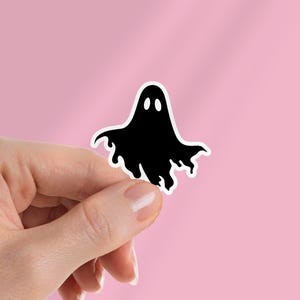 May include: A black ghost sticker with a white border. The ghost has two oval eyes and a flowing, wavy bottom edge. The sticker is held against a pink background.