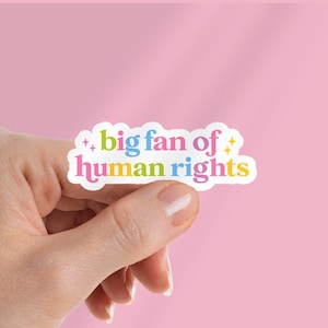 May include: A hand holds a white sticker with a scalloped edge. The sticker has the text "big fan of human rights" in colourful bubble letters. The background is a solid pink.