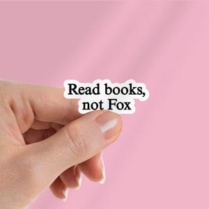 May include: A white sticker with the text "Read books, not Fox" in a black, sans-serif font. The sticker is held between fingers against a pink background.