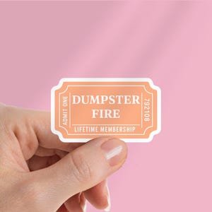 以下が含まれることがあります： 「DUMPSTER FIRE」と「LIFETIME MEMBERSHIP」の白い文字が入った、チケット型のピーチ色のステッカー。ステッカーには「ADMIT ONE」とシリアル番号も記載されています。背景は単色のピンク色です。