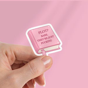 Op de afbeelding: Een roze sticker in de vorm van een boek met de tekst "PLOT? BABE, THEY'RE JUST FUCKING". De sticker heeft een witte rand en een roze lint-bladwijzer. De achtergrond is effen roze.