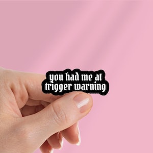 May include: A black sticker with the text "you had me at trigger warning" in a gothic font. The sticker is held by a hand against a pink background.