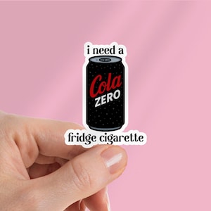 Puede incluir: Una pegatina con una lata negra de Cola Zero con el texto "Cola Zero" en rojo y blanco. La pegatina también tiene las frases "i need a" y "fridge cigarette" en texto blanco. La pegatina está sujeta por una mano sobre un fondo rosa.