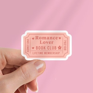 May include: A pink ticket-shaped sticker with the words "Romance Lover BOOK CLUB Lifetime Membership" in a serif font. The sticker has a light pink border and a white background. A hand holds the sticker against a pink background.