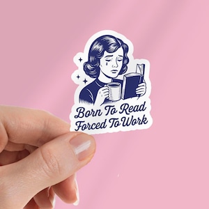 May include: A sticker with a vintage illustration of a woman with a tear, reading a book and holding a mug. The text on the sticker reads "Born To Read Forced To Work". The sticker is white with a blue illustration and is held by a hand against a pink background.
