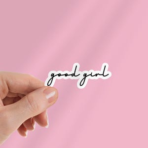 May include: A white sticker with the words "good girl" in a black, cursive font. The sticker is held between fingers against a pink background.