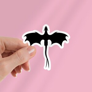 May include: A black dragon sticker with a white border. The dragon has outstretched wings and a long tail. The sticker is held against a pink background.