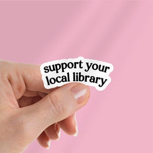Puede incluir: Una pegatina blanca con el texto "support your local library" en negro y negrita. La pegatina se presenta sobre un fondo rosa.