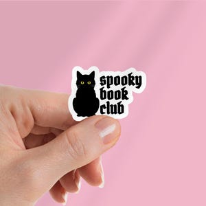 May include: A sticker featuring a black cat with yellow eyes and the words "spooky book club" in a gothic font. The cat is sitting upright. The sticker is held against a pink background.