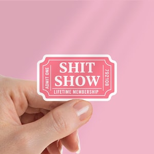 以下が含まれることがあります： 白い文字で「SHIT SHOW」と書かれたピンク色のチケット型のステッカー。ステッカーには「ADMIT ONE」と「LIFETIME MEMBERSHIP」の文字も含まれています。背景は単色のピンク色です。