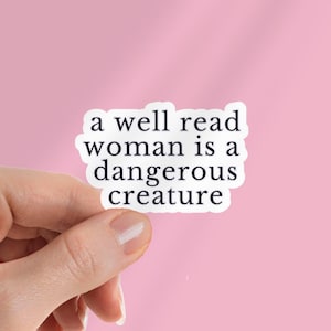 May include: A white sticker with the text "a well read woman is a dangerous creature" in a black, bold font. The sticker is held by a hand against a pink background. The sticker has a white border.