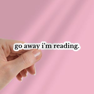 Könnte beinhalten: Ein weißer Aufkleber mit dem Text "go away i'm reading" in schwarzer, fetter Schrift. Der Aufkleber wird von einer Hand vor einem rosa Hintergrund gehalten. Der Aufkleber ist rechteckig mit abgerundeten Ecken.