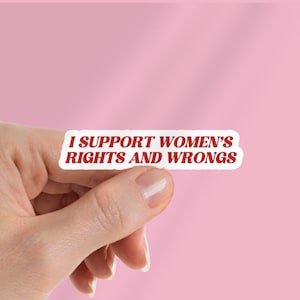 Puede incluir: Una pegatina blanca con texto rojo que dice "I SUPPORT WOMEN'S RIGHTS AND WRONGS" se sostiene sobre un fondo rosa. La pegatina es rectangular con esquinas redondeadas. El texto está en negrita, fuente sans-serif.