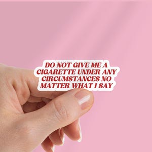 May include: A white sticker with red text that reads "DO NOT GIVE ME A CIGARETTE UNDER ANY CIRCUMSTANCES NO MATTER WHAT I SAY" is held against a pink background. The sticker is in a rounded rectangle shape.