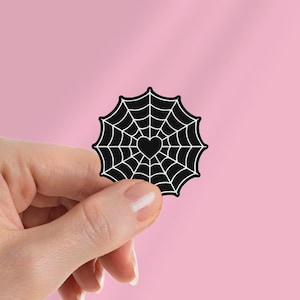 May include: A black and white sticker with a spiderweb design and a heart at the centre. The sticker is held against a pink background. The design has a gothic or Halloween theme.