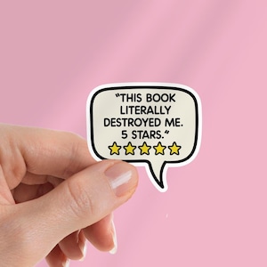 May include: A white sticker in the shape of a speech bubble with the text "THIS BOOK LITERALLY DESTROYED ME. 5 STARS." and five gold stars. The sticker is held against a pink background.