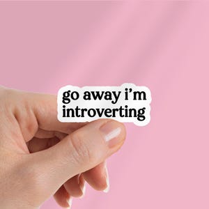 Puede incluir: Una pegatina blanca con la frase "go away i'm introverting" en texto negro. La pegatina está sujeta por una mano sobre un fondo rosa.