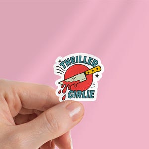 May include: A sticker with a cartoon knife design. The knife is positioned over a red circle, with blood-like drips. The text "THRILLER GIRLIE" is included in the design. The sticker is held in a hand against a pink background.