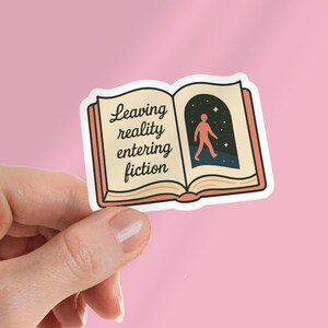 May include: A sticker featuring an open book with the text "Leaving reality entering fiction." The right page shows a person walking into a starry night scene. The book has a cream-colored interior and a pink border.
