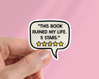 Könnte beinhalten: Ein weißer Aufkleber in Form einer Sprechblase mit dem Text "THIS BOOK RUINED MY LIFE. 5 STARS." und fünf goldenen Sternen. Der Aufkleber wird vor einem rosa Hintergrund gehalten.