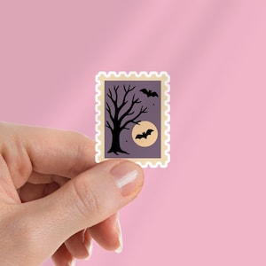 May include: A Halloween-themed sticker featuring a spooky tree silhouette, bats, and a full moon. The sticker has a purple background and a cream-colored border with a postage stamp design. The sticker is held by a hand against a pink background.
