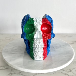 May include: A white sugar skull with blue, green, and red floral designs. The skull is painted in the colors of the Italian flag.
