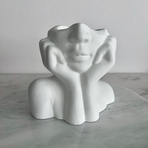 May include: White ceramic planter shaped like a woman's bust with her hands resting on her chin. The planter has a hollow space in the top of the head for planting.