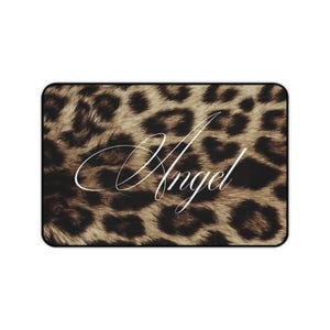Peut inclure: Un tapis rectangulaire avec un motif imprimé léopard marron et noir. Le mot Angel est écrit au centre dans une police cursive blanche. Le tapis présente une fine bordure noire qui encadre l'ensemble du motif imprimé léopard.