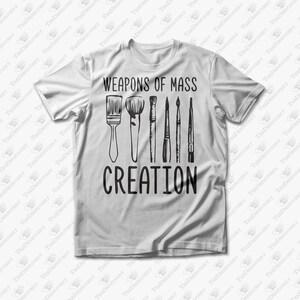 May include: White t-shirt featuring the text "WEAPONS OF MASS CREATION" above a graphic of paintbrushes. The design is in black and is centered on the front of the shirt. The shirt is a short-sleeved crew neck.