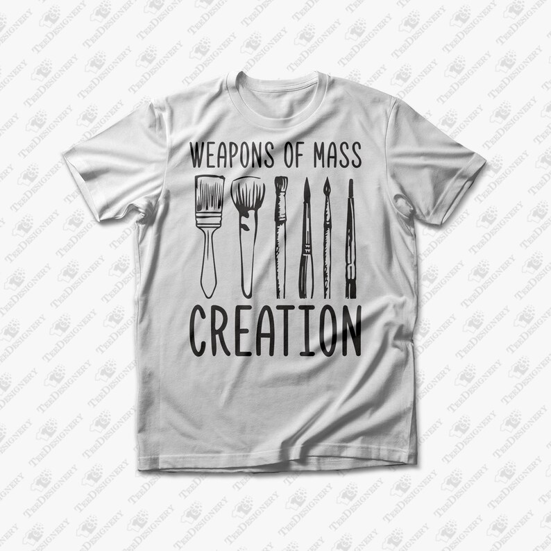 May include: White t-shirt featuring the text "WEAPONS OF MASS CREATION" above a graphic of paintbrushes. The design is in black and is centered on the front of the shirt. The shirt is a short-sleeved crew neck.