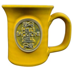 Puede incluir: Taza de cerámica amarilla con asa curva. La taza presenta un emblema dorado con el texto "HELL'S BACKBONE GRILL BOULDER, UTAH" en un diseño circular. La taza tiene una parte superior acampanada y un acabado brillante.