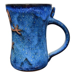 May include: A handmade ceramic mug with a blue glaze and brown starfish designs. The mug has a large handle and a speckled texture. The mug is approximately 4 inches tall.