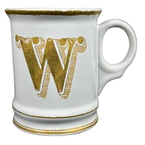 May include: White ceramic mug featuring a gold-coloured letter "W". The mug has a rounded handle and gold trim around the top and bottom. The letter "W" is in an ornate font, adding a touch of elegance.