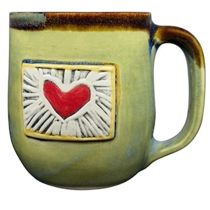 Może przedstawiać: Ręcznie robiony ceramiczny kubek z jasnozieloną glazurą. Kubek posiada wypukły prostokątny wzór z czerwonym sercem pośrodku, otoczonym białymi promieniami. Obręcz i uchwyt mają brązową glazurę.