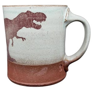 May include: A handmade ceramic mug with a dinosaur silhouette. The mug features a light blue and brown color scheme, with a reddish-brown Tyrannosaurus Rex design on the side. The handle is white.