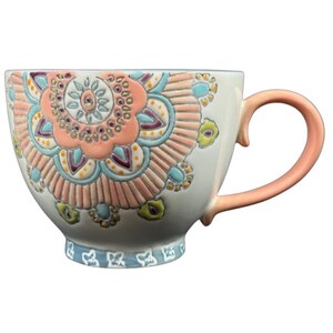 May include: A large, white ceramic mug with a decorative floral design in shades of peach, blue, and yellow. The handle and the center of the floral design are peach-colored. The base of the mug has a blue patterned border.