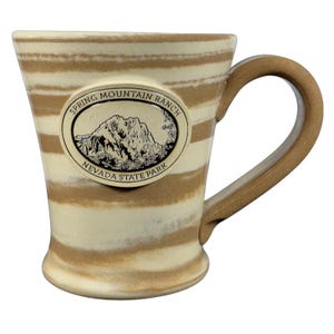 Op de afbeelding: Een keramische mok met een beige en bruin gestreept patroon. De mok heeft een gebogen handvat en een ovaal embleem met de tekst "Spring Mountain Ranch Nevada State Park" en een bergillustratie.
