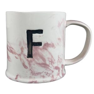 Può includere: Tazza in ceramica bianca con un design marmorizzato rosa e la lettera nera "F". La tazza ha un manico curvo e una base leggermente svasata. La marmorizzazione rosa crea un motivo unico sulla superficie della tazza.