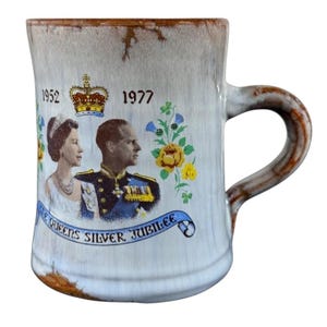 May include: A vintage ceramic mug commemorating the Queen's Silver Jubilee, featuring portraits of the Queen and Prince Philip, with the years 1952 and 1977. The mug has a light blue and brown color scheme with a decorative handle and floral accents.